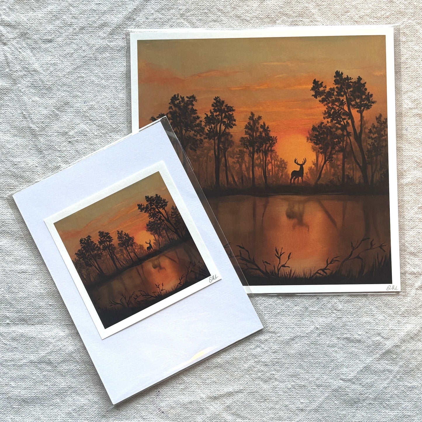 Two prints of a sunset over a lake with trees and a deer, one inside a white frame on a textured surface.