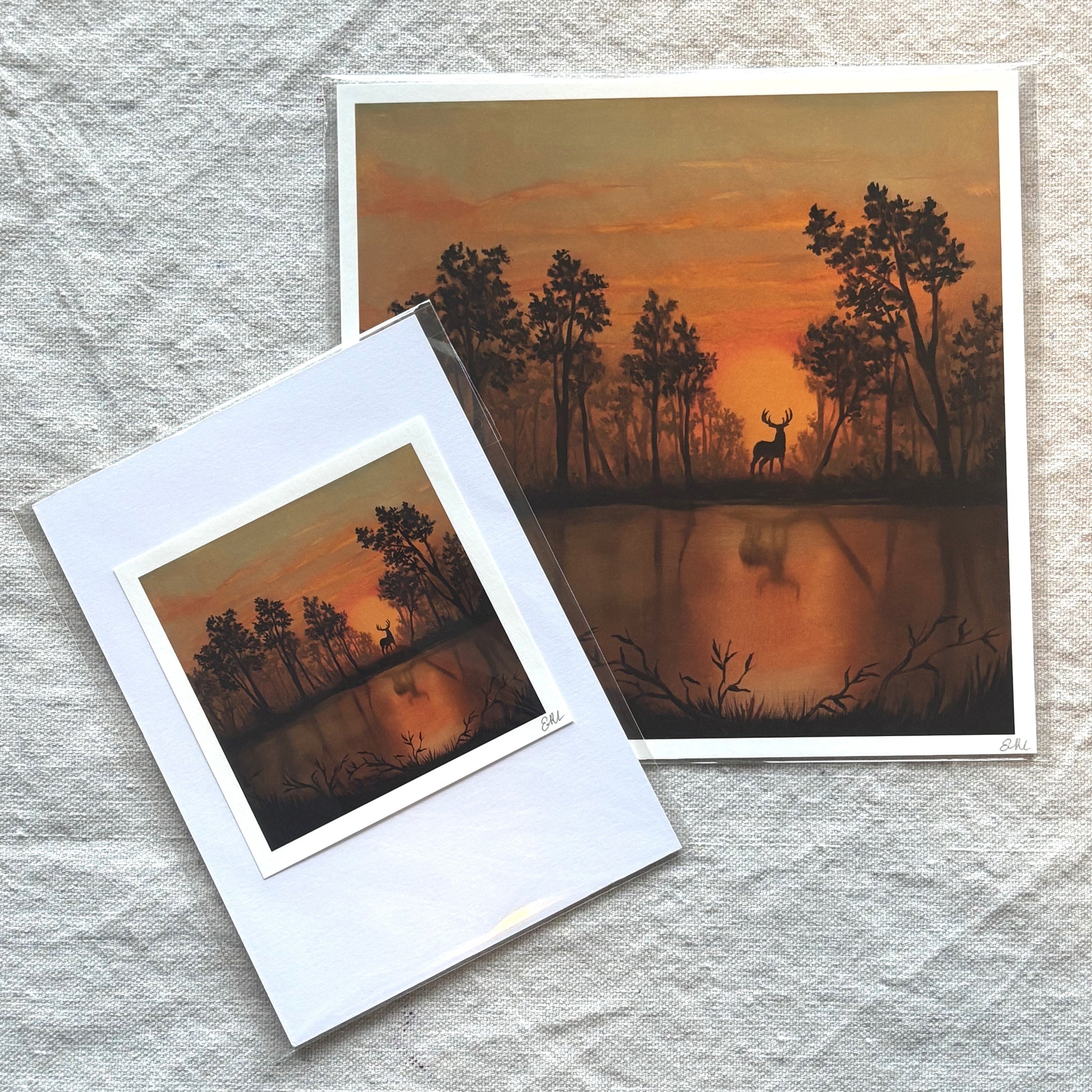 Two prints of a sunset over a lake with trees and a deer, one inside a white frame on a textured surface.