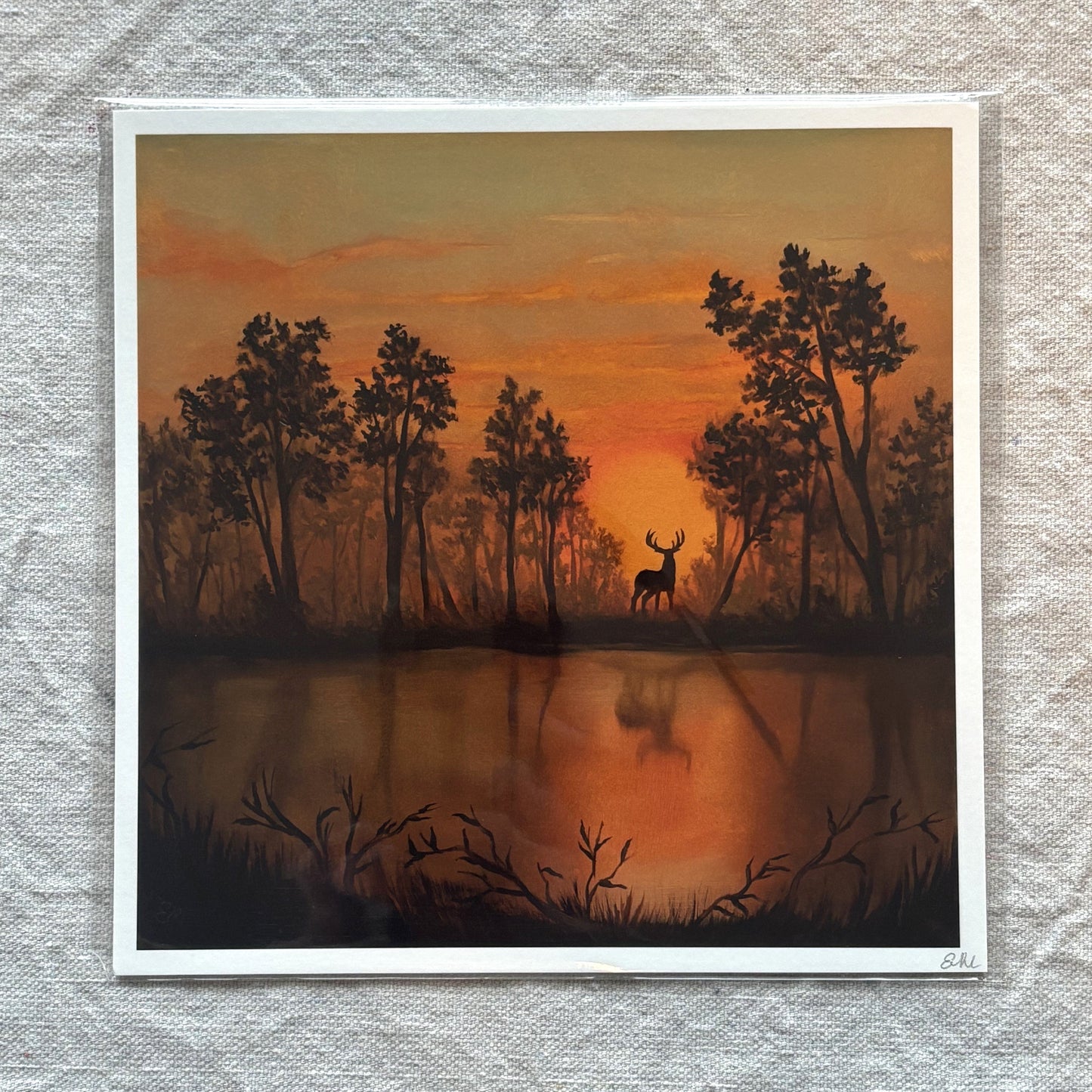 art print of a deer silhouette at sunset over water with trees, framed on a textured wall.