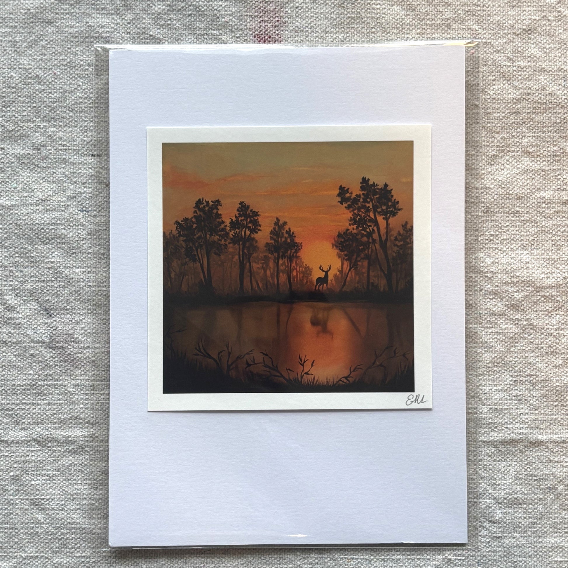 Art print of a deer by a lake with trees at sunset, framed and on a textured fabric background.