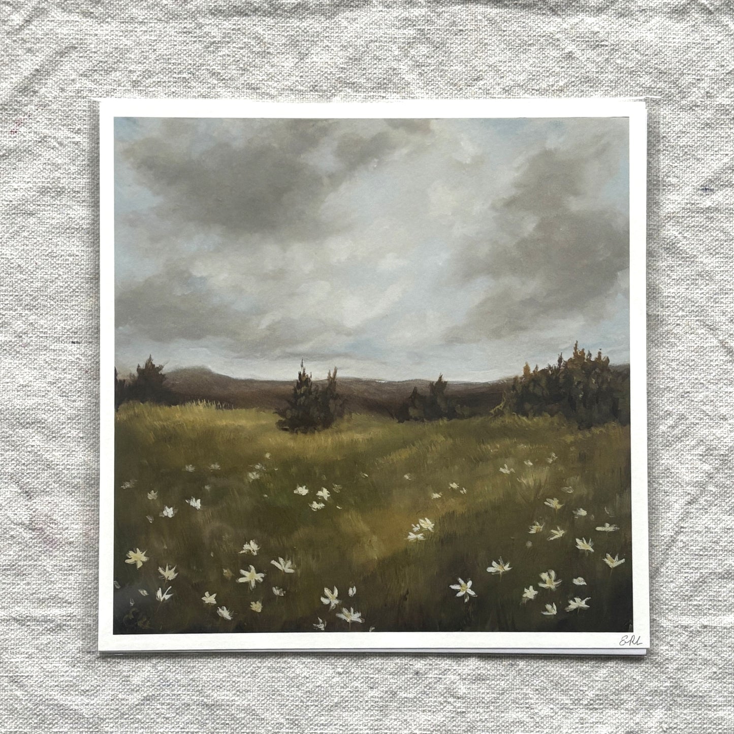 Print of a landscape with a field, trees, and a cloudy sky on a textured gray background.
