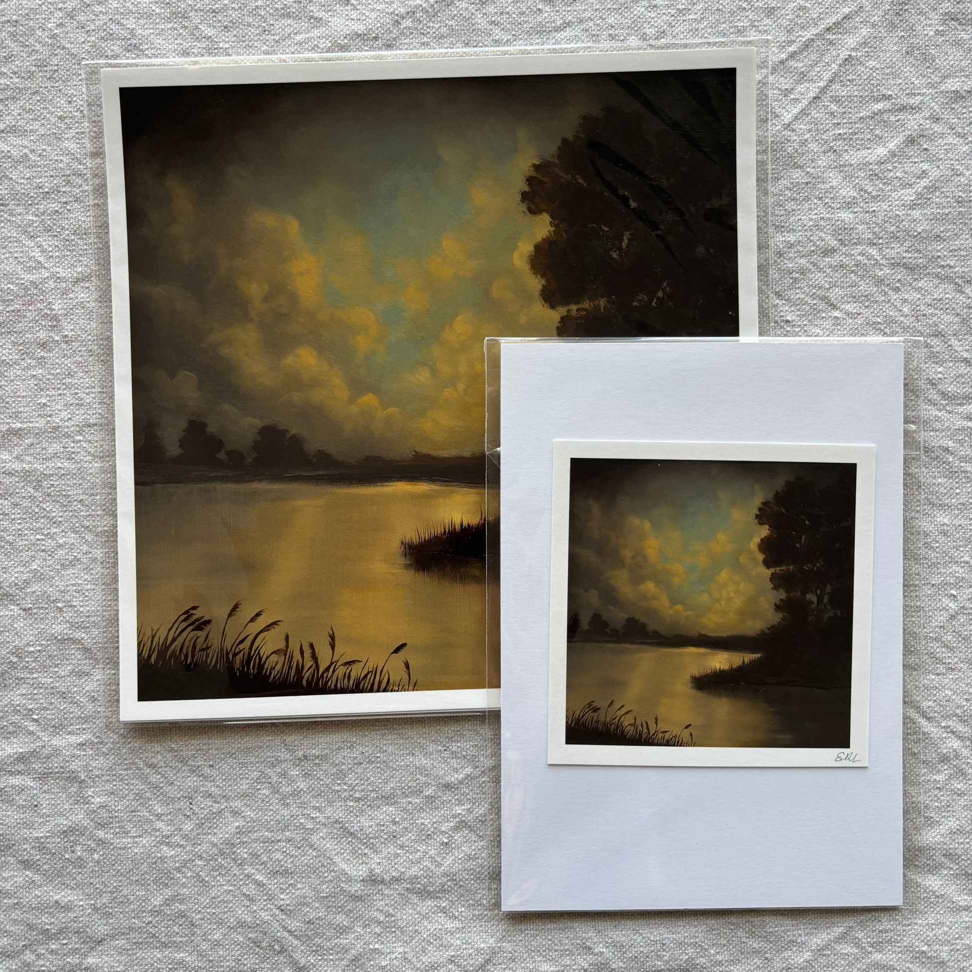 Two art prints of a landscape scene with a lake and trees on a textured gray surface.