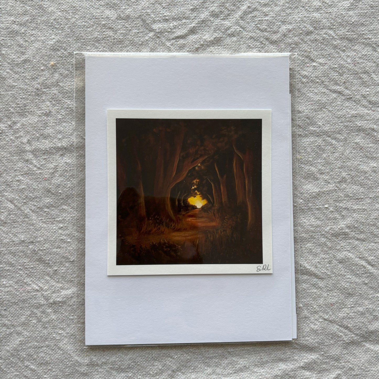 Small art print of a forest scene with a glowing object, framed by a white border on a textured gray surface.